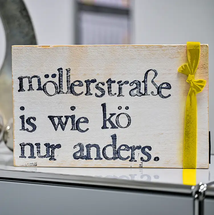 Ein Holzschild mit pinker Schleife und der Aufschrift "Möllerstraße ist wie kö, nur anders".